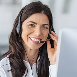 Smiling woman with long dark hair wearing a headset, sitting at a desk and looking at a computer screen, appearing to provide customer service or technical support.