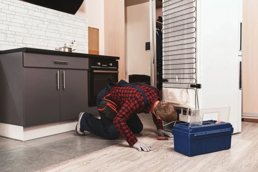 A technician in work overalls and gloves kneels on a kitchen floor, inspecting the back of a refrigerator with tools from an open blue toolbox beside him. The kitchen features dark cabinets and light wood flooring.
