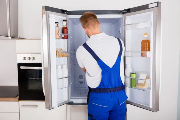 A repairman in blue overalls stands with his back to the camera, working on an open refrigerator in a modern kitchen. Various food items and bottles are visible inside the fridge.