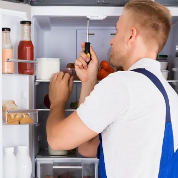 A repairman in a white shirt and blue overalls uses a screwdriver to fix the inside of a refrigerator, with food items and bottles visible on the shelves.