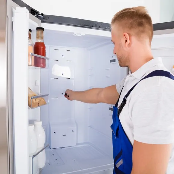 A man in blue overalls uses a screwdriver to repair the inside of an open refrigerator that contains a few bottles and cartons on the shelves.