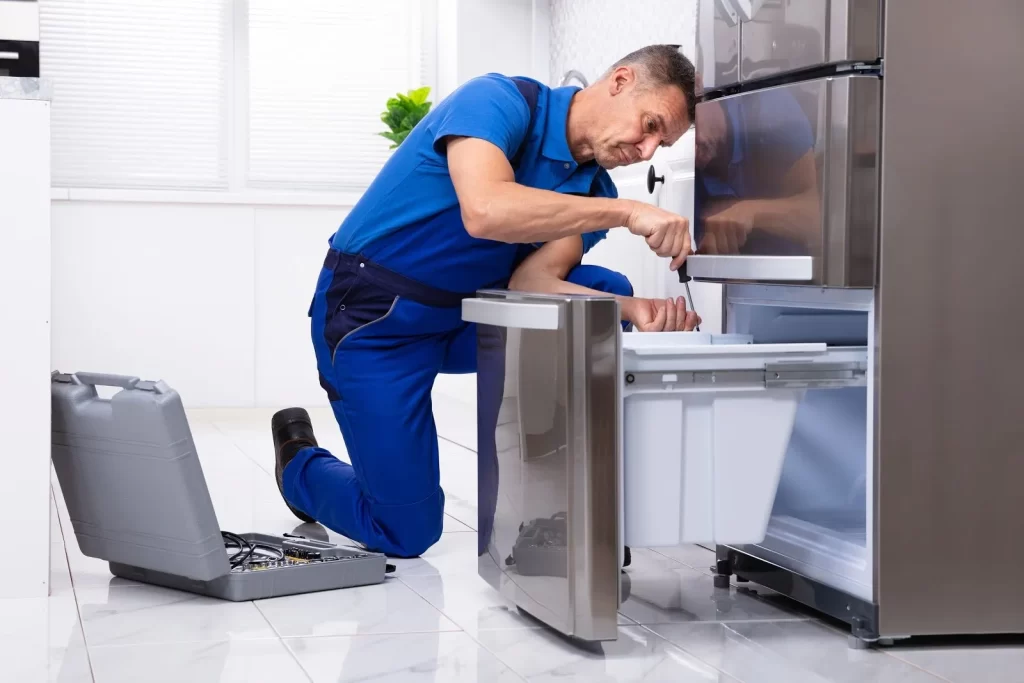 A repairman in blue overalls kneels on a kitchen floor, fixing the open lower drawer of a stainless steel refrigerator, with a toolbox and tools beside him.