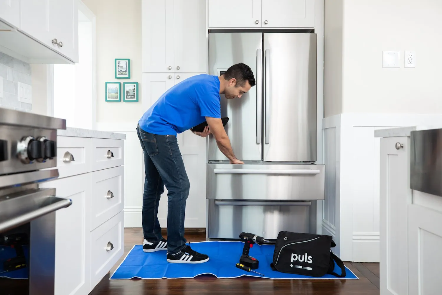 A repair technician in a blue shirt inspects the bottom freezer drawer of a stainless steel refrigerator in a modern kitchen. Tools and a black Puls bag are on a blue mat on the floor nearby.