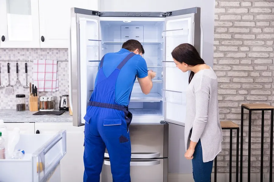 A repair technician in blue overalls examines the inside of an open refrigerator while a woman stands nearby, watching attentively in a modern kitchen.
